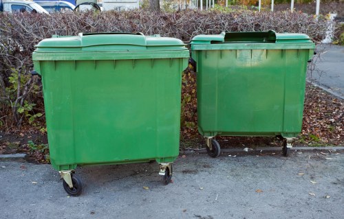 Two large green wheeled rubbish bins positioned on a paved surface outdoors, with the left bin being slightly larger than the right. The bins are made of solid plastic with a textured finish and feature hinged lids on top. The larger bin appears to be empty, while the smaller one has a few faint marks on its surface. They are located on a concrete or asphalt area, possibly a driveway or parking space, with sparse shrubbery and a fence visible in the background. The scene is evenly lit, suggesting daytime with natural light, and the bins are situated in an area that might be used for waste collection in an urban or suburban setting. These types of rubbish bins are commonly used for general waste disposal in commercial or residential premises, such as offices or communal areas, and the image relates to waste management services like those offered by Office Clearance Elephant and Castle, serving local regions around the postcode and town in central London.