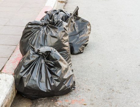 The image shows two large black rubbish bags made of plastic, filled with waste material, placed on the edge of a concrete pavement next to a curb on an urban street. The bags appear to be tightly sealed and bulging with their contents, which are not visible. They are positioned close to a painted red line along the curb, indicating a designated parking or loading area, with a section of worn, light-colored pavement visible to the left. The street surface is smooth and grey, with some scattered small debris nearby. The surroundings suggest a typical urban environment suitable for rubbish removal services such as Office Clearance Elephant and Castle, which operates in the local postcode area. The lighting is natural, indicating daytime conditions, and the scene reflects general waste collection or rubbish disposal activity typical in a residential or commercial street setting in the vicinity of Elephant and Castle.
