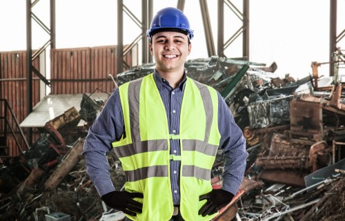 A male waste management worker inside a large industrial warehouse, standing in front of a pile of mixed construction and commercial waste materials including broken wood, metal, and plastic debris. He is wearing a blue safety helmet, a high-visibility yellow vest over a blue shirt, and black gloves, smiling confidently at the camera. The background shows metal framework and windows, with natural light illuminating the scene, emphasizing the context of rubbish removal services provided by Office Clearance Elephant and Castle in the local area. The debris appears to be part of a recent clearance project, and the environment reflects a professional setting focused on waste collection and recycling efforts.