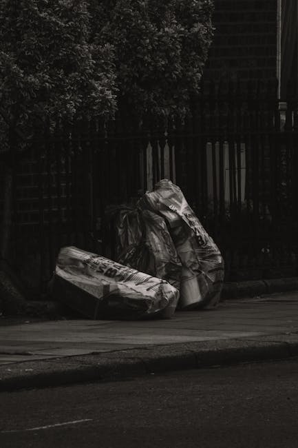 A large collection of overflowing rubbish and waste materials is gathered on a paved urban sidewalk, positioned near a metal railing that surrounds a small parking area. The waste includes a variety o