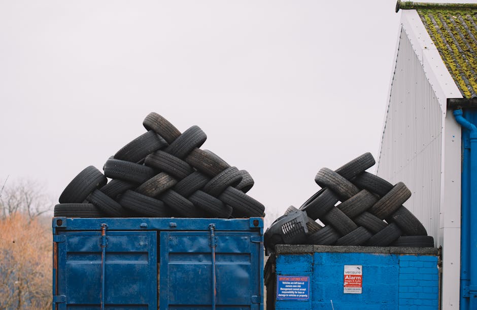 A large heap of mixed metallic and plastic waste consisting of discarded appliances, metal frames, and broken furniture, piled up outdoors against a background of a clear blue sky. The materials displ