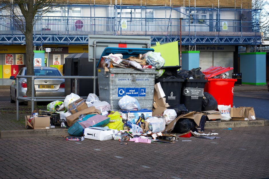 A large metal waste collection bin situated on a paved urban street, overflowing with various types of rubbish including cardboard boxes, plastic bags, and paper, with some waste spilling onto the sur