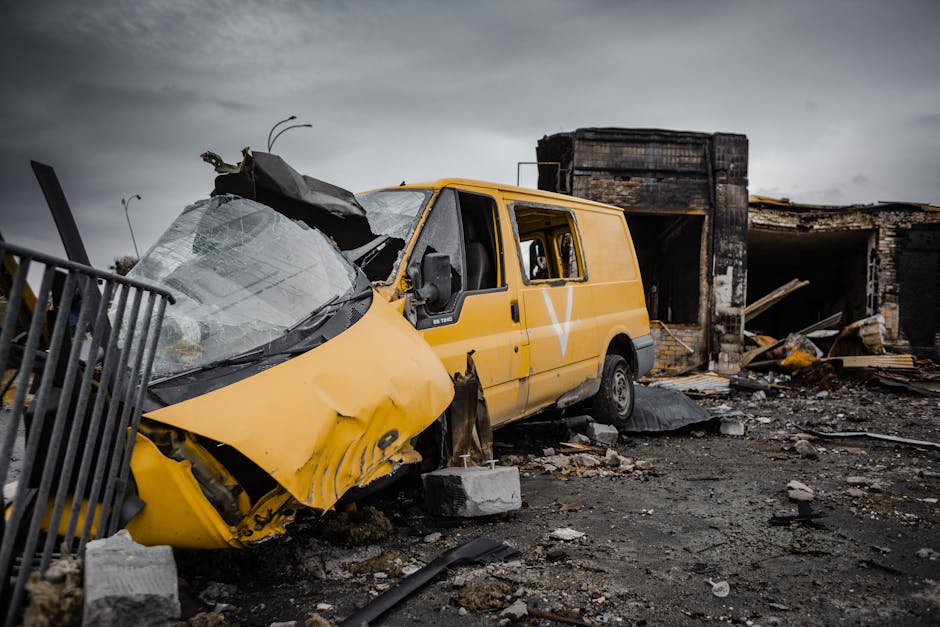 A yellow utility van with significant front-end damage, including a crushed hood and shattered windshield, is situated on a debris-strewn ground near a partially collapsed brick building. The van's ri